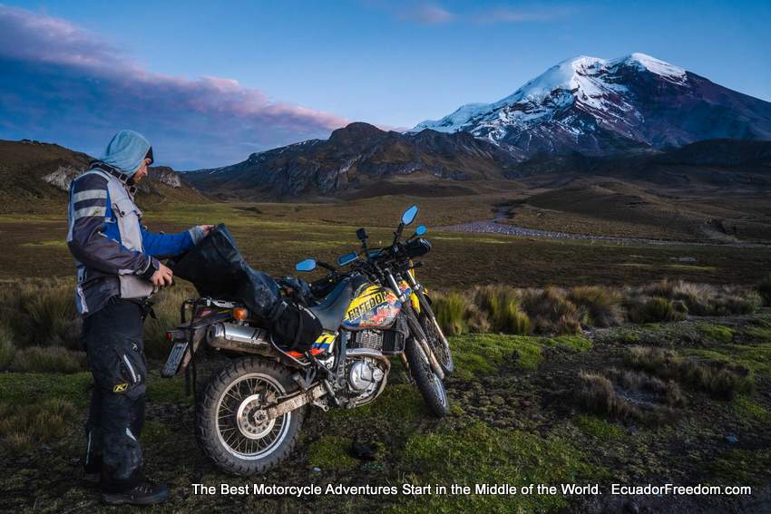 adventure motorcyclist looking into a saddlebag with snow covered mountain in the background